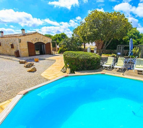 Foto desde la piscina con las hamacas en el borde y la villa de estilo rústico al fondo rodeada de jardines en Villa Tres Pinos.